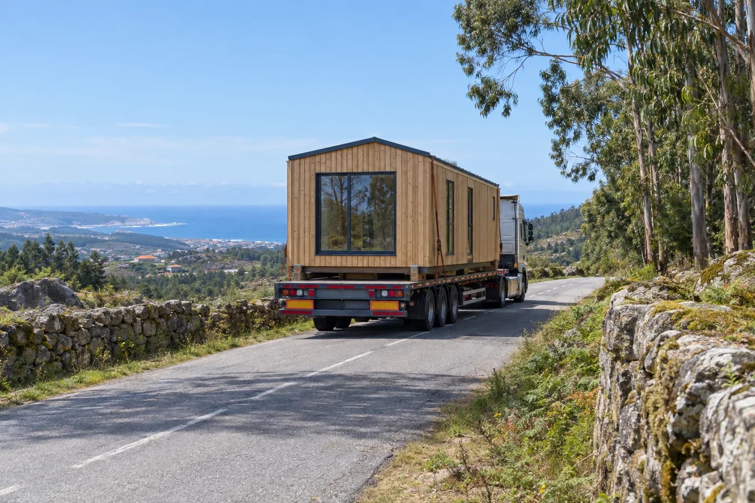 Camião de caixa aberta a transportar um módulo de casa de madeira por uma estrada rural, com muros de pedra