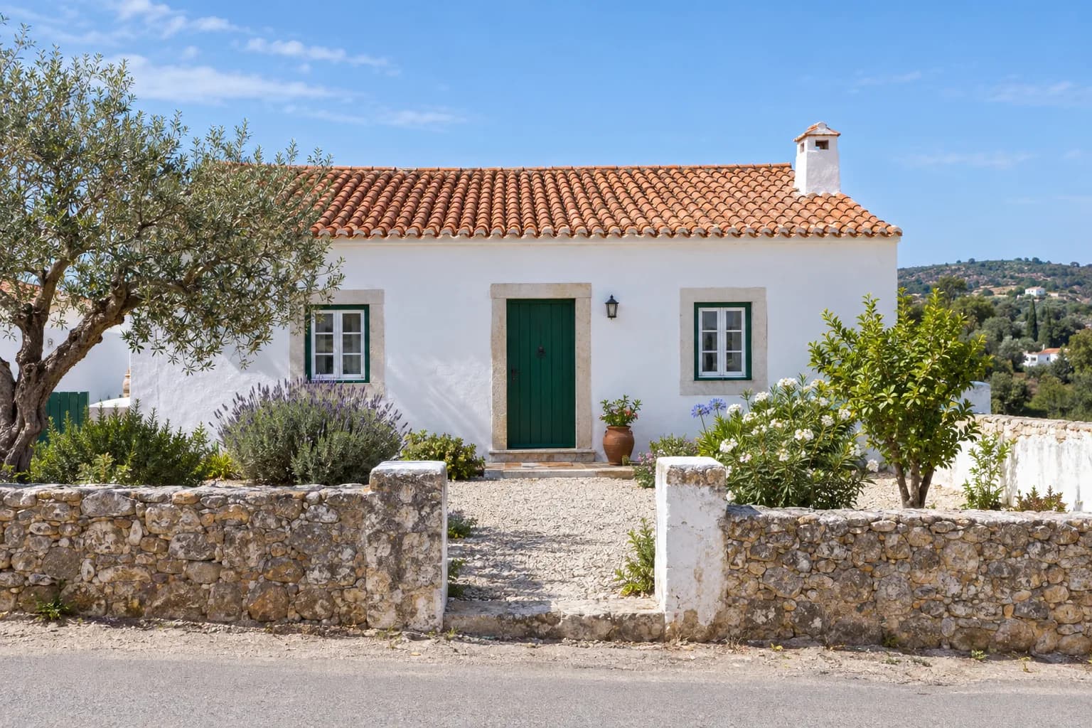 Casa branca térrea com telhado de telha e porta verde, vista da rua, com muro de pedra e uma oliveira